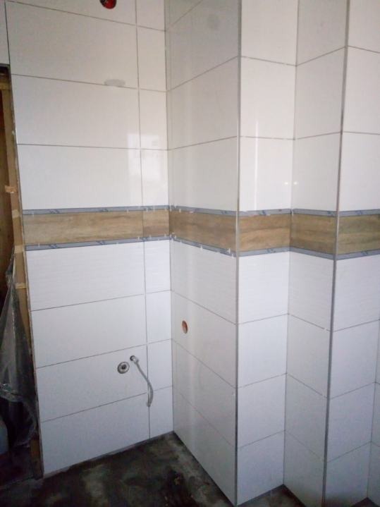 Ściany wyłożone białymi, poziomymi płytkami z dekoracyjnym, drewnopodobnym pasem, widoczny fragment instalacji wodnej i surowa posadzka.