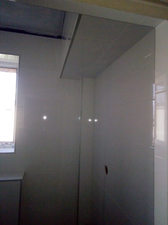 Wykończona łazienka z błyszczącymi, jasnymi płytkami ceramicznymi na ścianach i suficie; widoczne okno i wystający element sufitu.