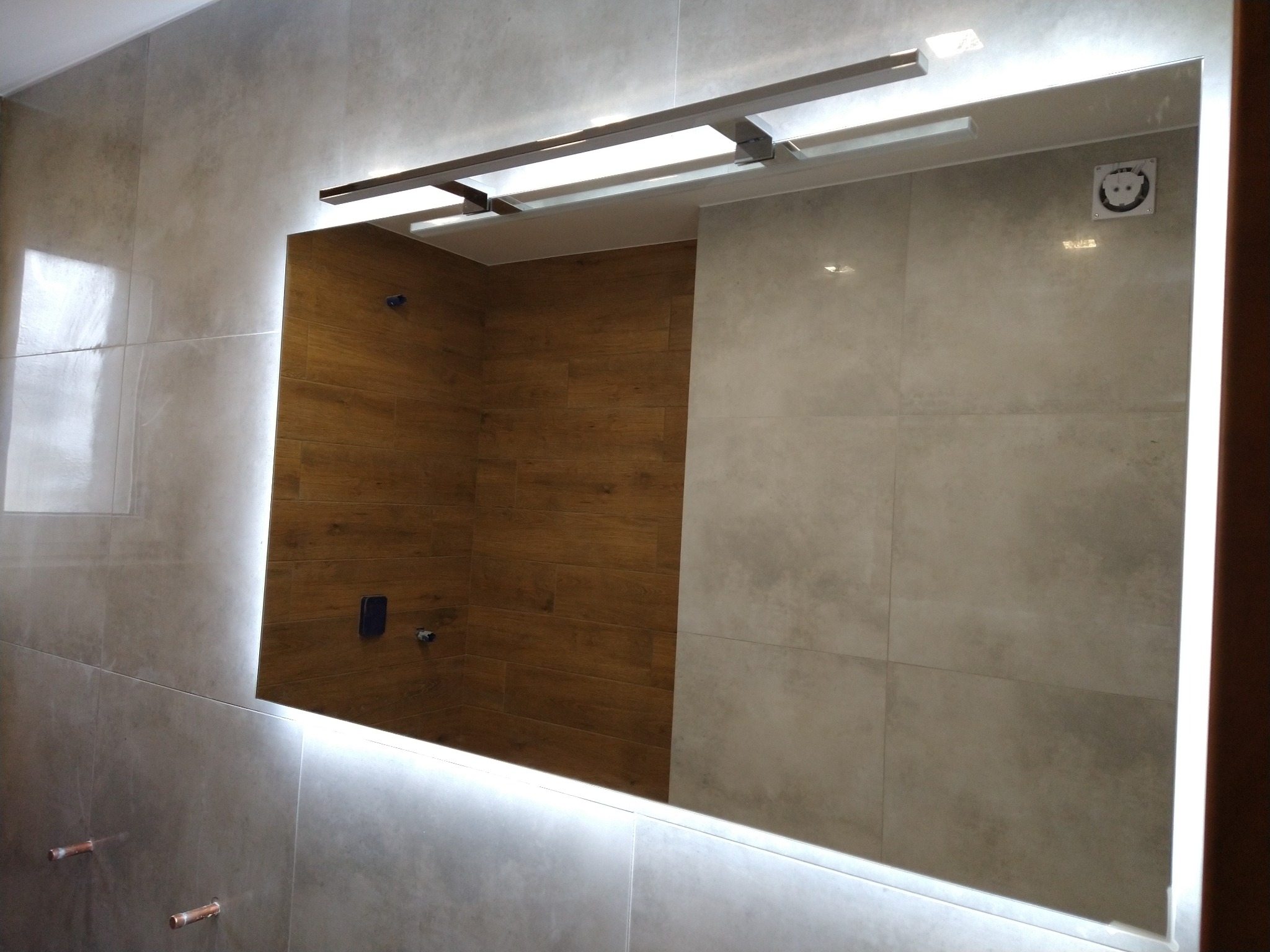 Nowoczesna łazienka z podświetlanym lustrem, widoczne przyłącza wodne w ścianie oraz gniazdko elektryczne.