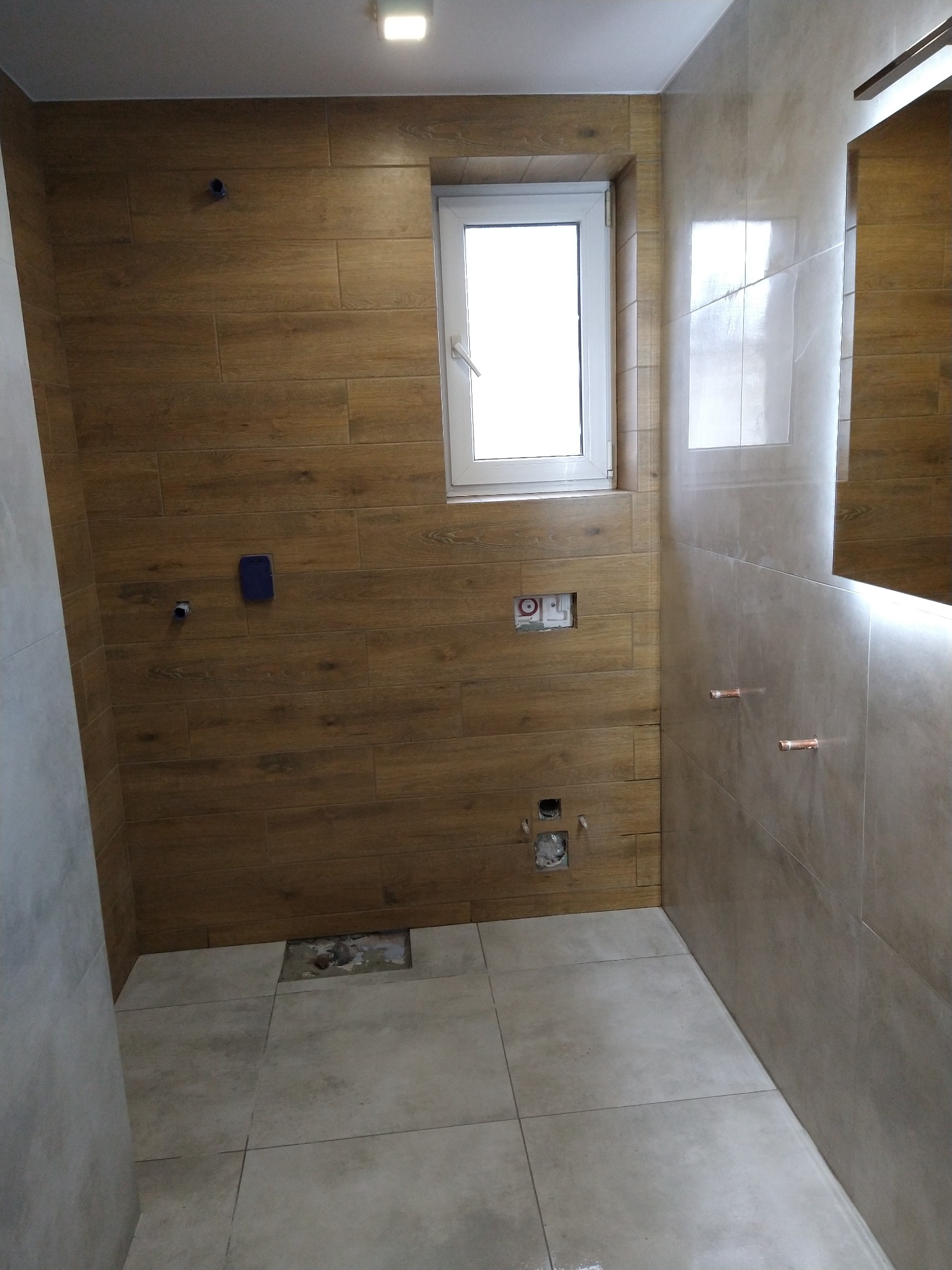 Remontowana łazienka z drewnopodobnymi płytkami ściennymi, widoczne otwory na instalacje, brakująca płytka podłogowa i białe okno.