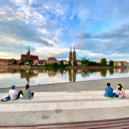 Panorama Wrocławia z Ostrowem Tumskim odbijająca się w wodach Odry; grupa turystów siedzi na nabrzeżu, podziwiając widok w słoneczny dzień.
