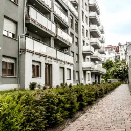 Nowoczesny budynek apartamentowy z balkonami, widok z poziomu chodnika wyłożonego kostką brukową, zieleń wzdłuż ścieżki.