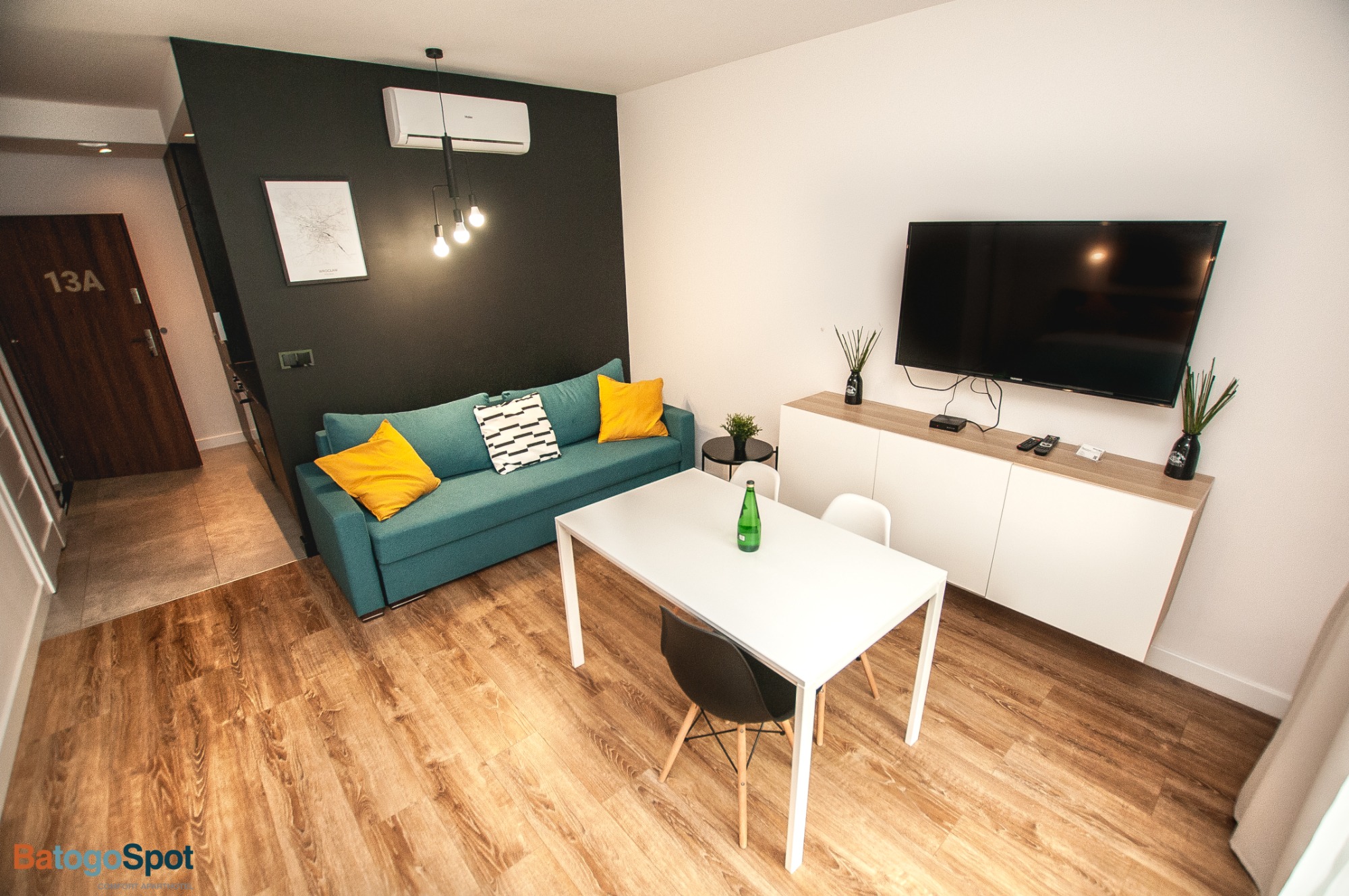 Nowoczesny pokój z turkusową sofą, żółtymi poduszkami, białym stołem i telewizorem na ścianie, oświetlony wiszącymi lampami i klimatyzacją.