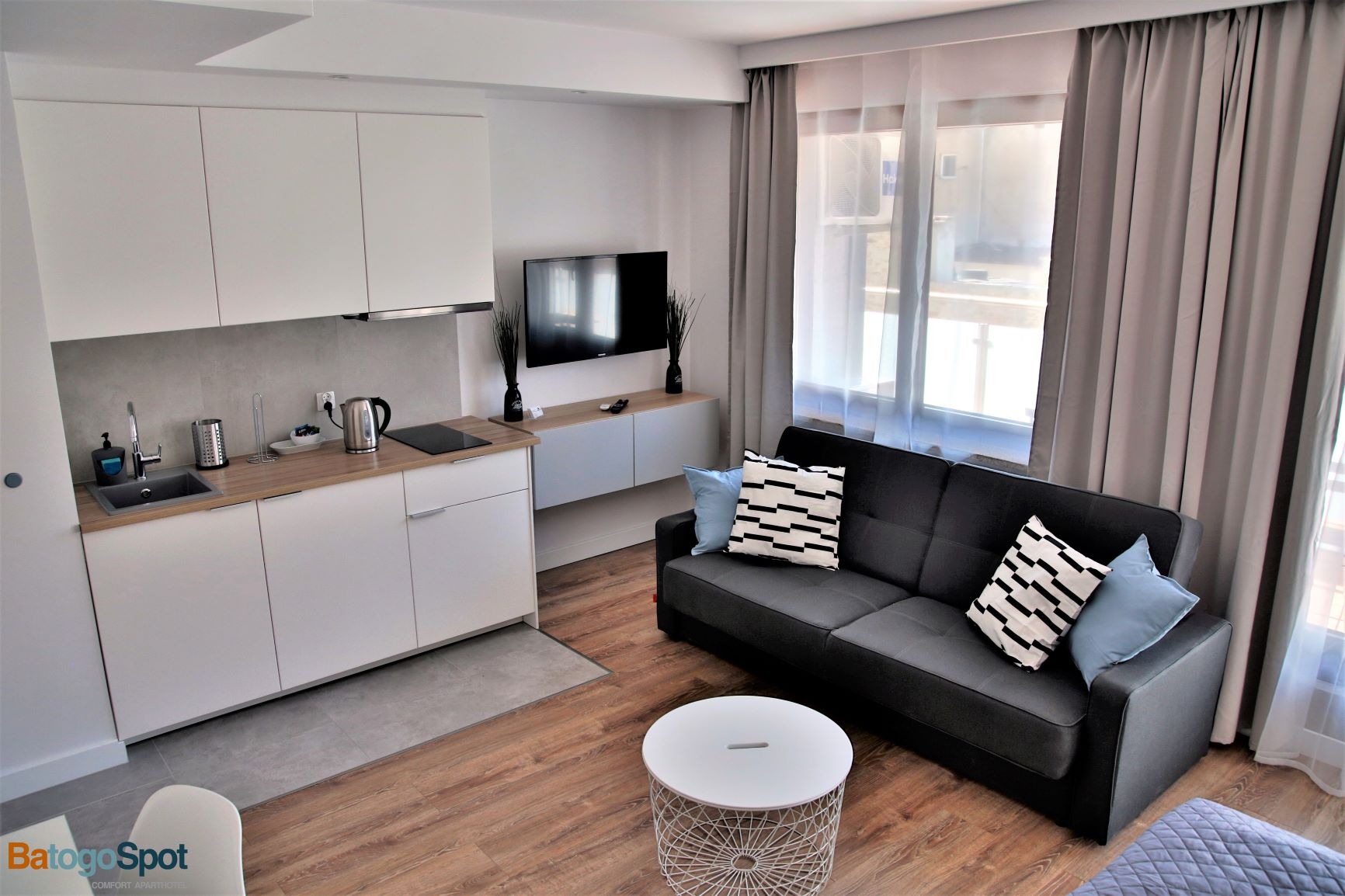 Nowoczesny apartament z aneksem kuchennym, szarą sofą z dekoracyjnymi poduszkami i okrągłym stolikiem kawowym na drewnianej podłodze.