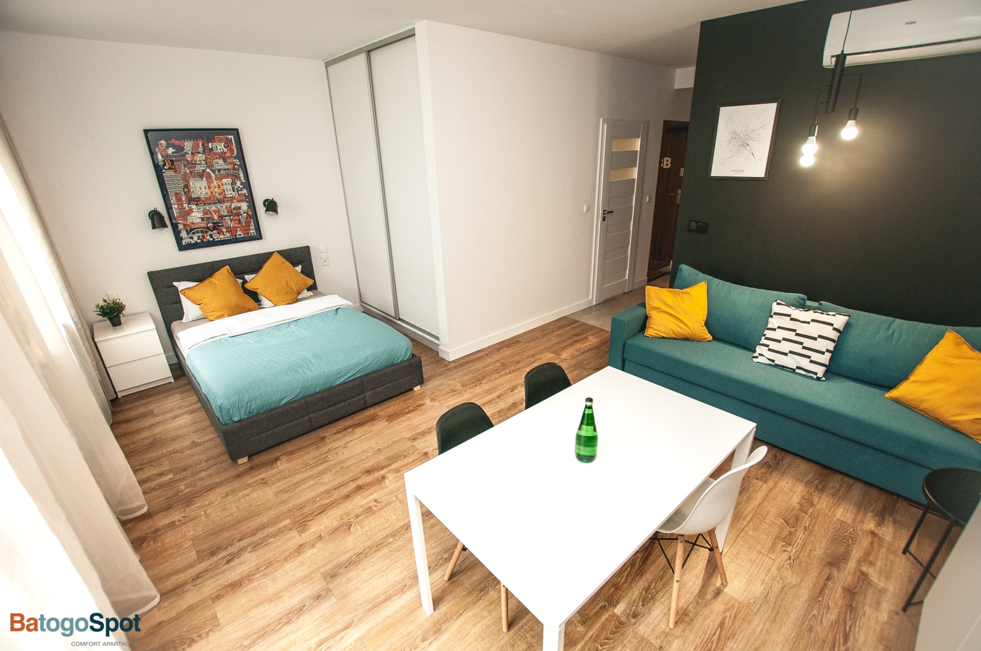 Jasne studio z łóżkiem, turkusową pościelą i żółtymi poduszkami, białym stołem z butelką wody, turkusową sofą z żółtymi poduszkami, drewnianą podłogą i czarną ścianą z klimatyzacją.