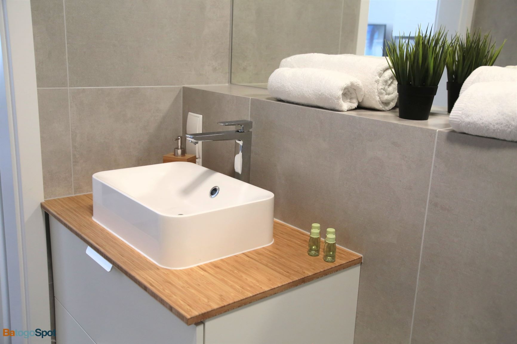 Nowoczesna łazienka z białą, prostokątną umywalką nablatową na bambusowym blacie, minimalistyczna bateria, dwie małe buteleczki kosmetyków i zwinięte białe ręczniki na tle szarych płytek.