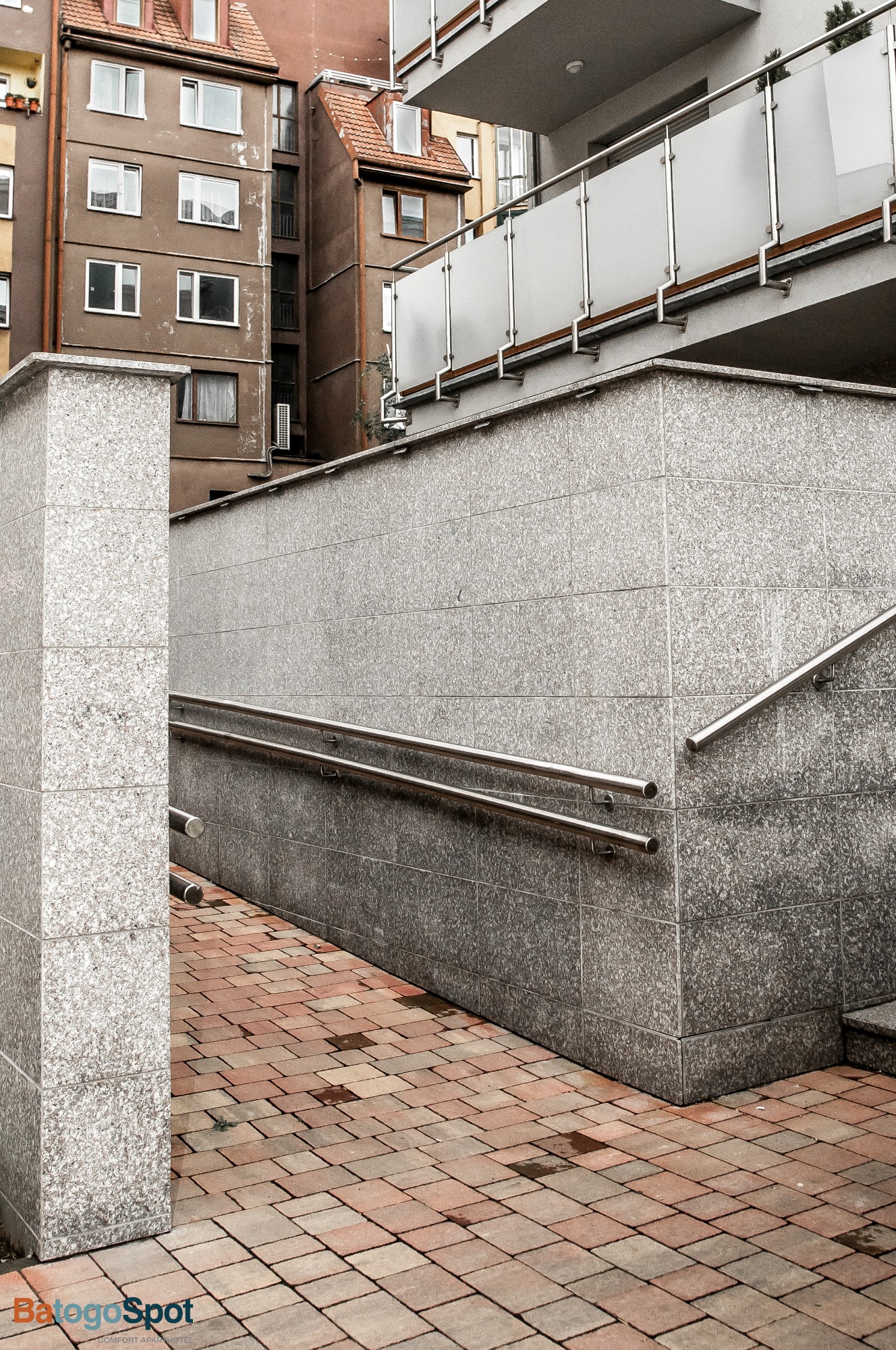 Zewnętrzne schody z podjazdem dla wózków, wyłożone szarą, nakrapianą płytką, z metalowymi poręczami i czerwoną kostką brukową na chodniku, na tle budynków mieszkalnych.