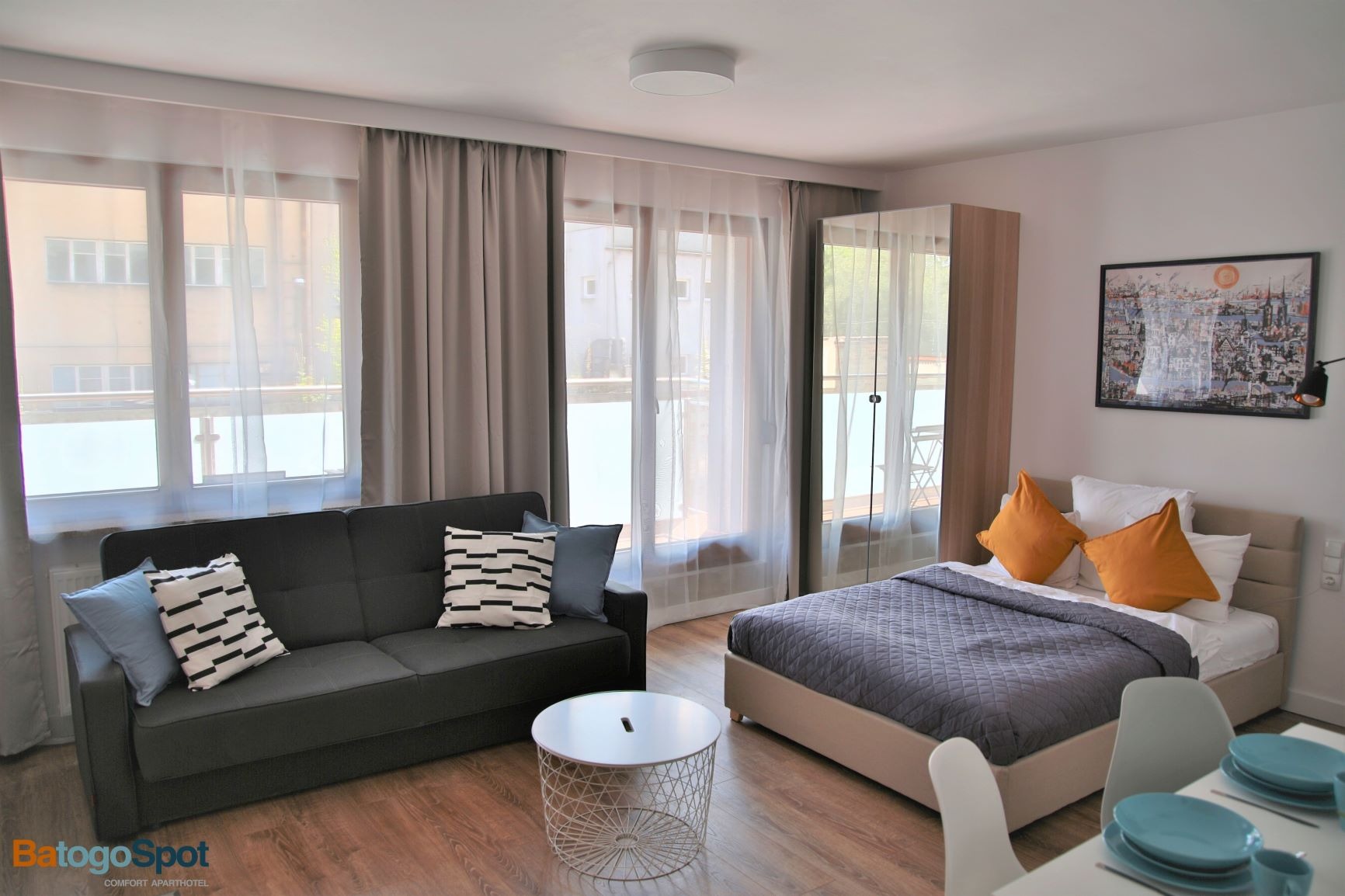 Jasne wnętrze apartamentu z szarym narożnikiem, okrągłym stolikiem kawowym z metalową podstawą, łóżkiem z szarą narzutą i żółtymi poduszkami oraz stołem z błękitną zastawą.