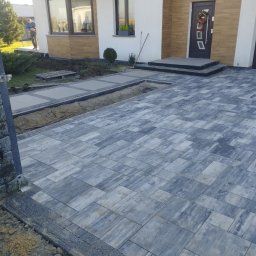 PBB DARKBRUK - Ułożona szara kostka brukowa przed domem z widocznym fragmentem przygotowanego podłoża i pracownikiem w tle. Wieniec świąteczny na drzwiach.