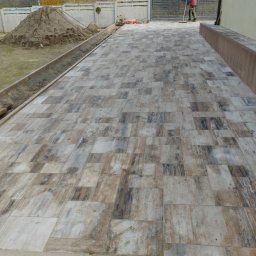PBB DARKBRUK - Ułożona kostka brukowa w odcieniach szarości i brązu tworzy wzorzysty chodnik. W tle widoczny stos piasku i pracownik z miotłą.