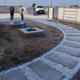 PBB DARKBRUK - Układanie ścieżki z płyt betonowych i białego grysu. W tle dwaj mężczyźni i brama wjazdowa. Widoczne studzienki kanalizacyjne z niebieskimi rurami.