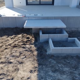 PBB DARKBRUK - Budowa elementu z kostki brukowej przy tarasie domu. Widoczny poziomica, świeży montaż z użyciem silikonu. Etap prac ziemnych.