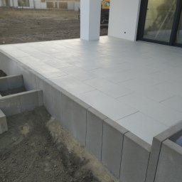 PBB DARKBRUK - Jasnoszara, prostokątna kostka na tarasie domu jednorodzinnego. Widoczne obrzeża i fragment terenu przed budynkiem. Minimalistyczny styl.