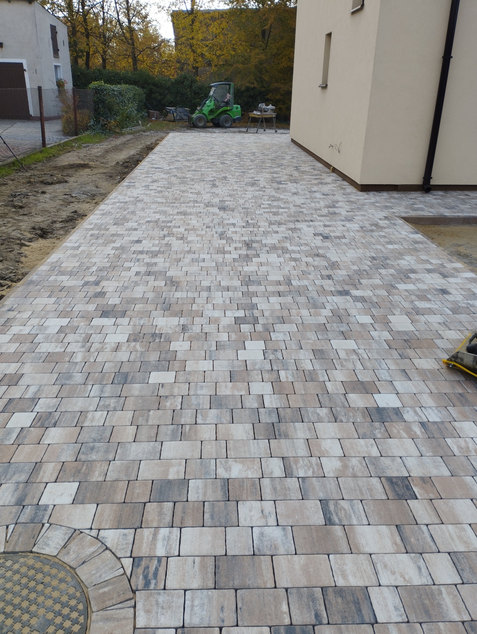 Ułożona kostka brukowa w odcieniach szarości i beżu, z widocznym włazem kanalizacyjnym na skraju, obok budynku z czarną rynną i zielonym miniładowarką w tle.