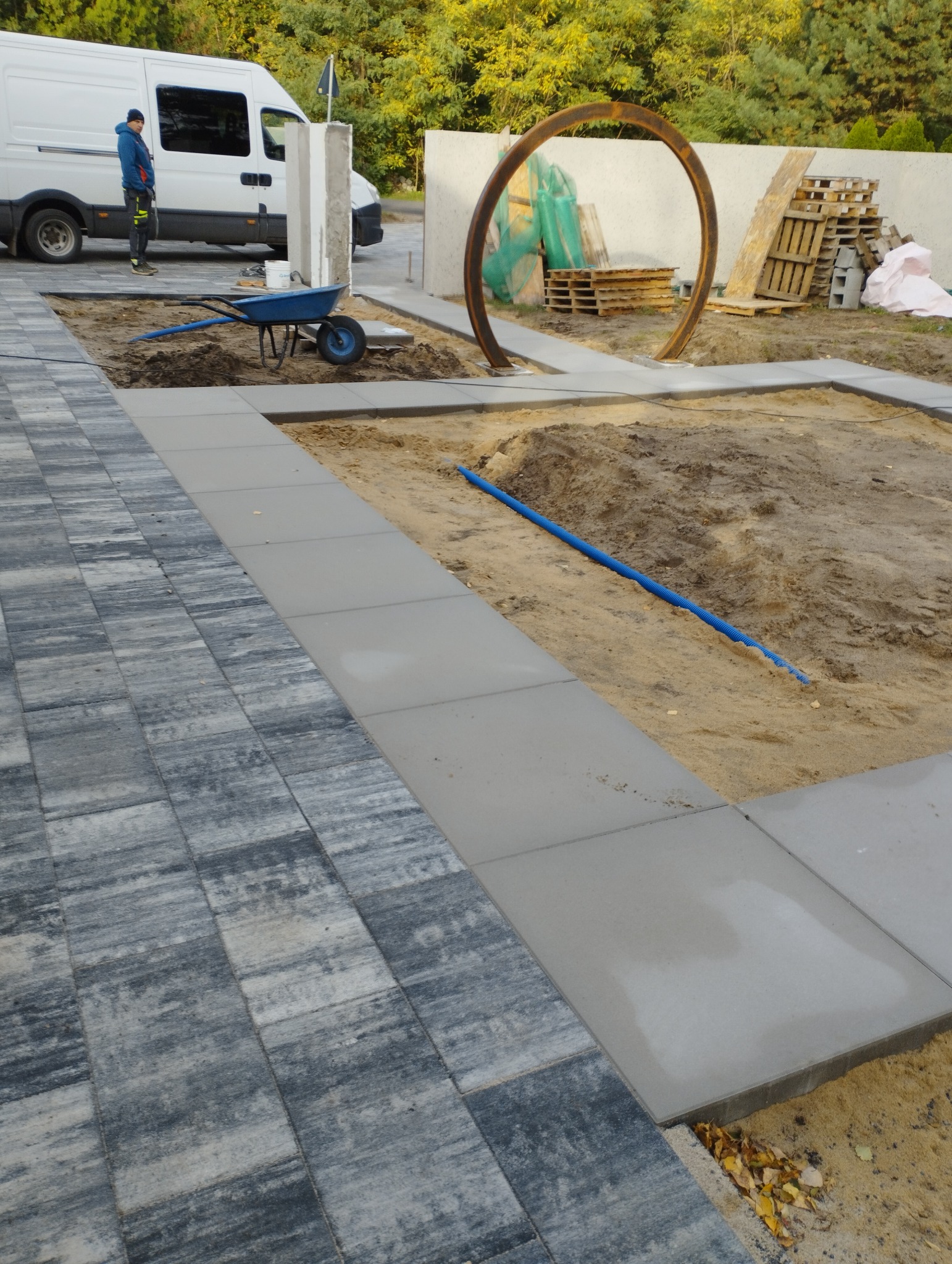 Fragment układanej nawierzchni z kostki brukowej i płyt betonowych, widoczna taczka z narzędziami, pracownik w tle i element dekoracyjny w postaci metalowego okręgu.