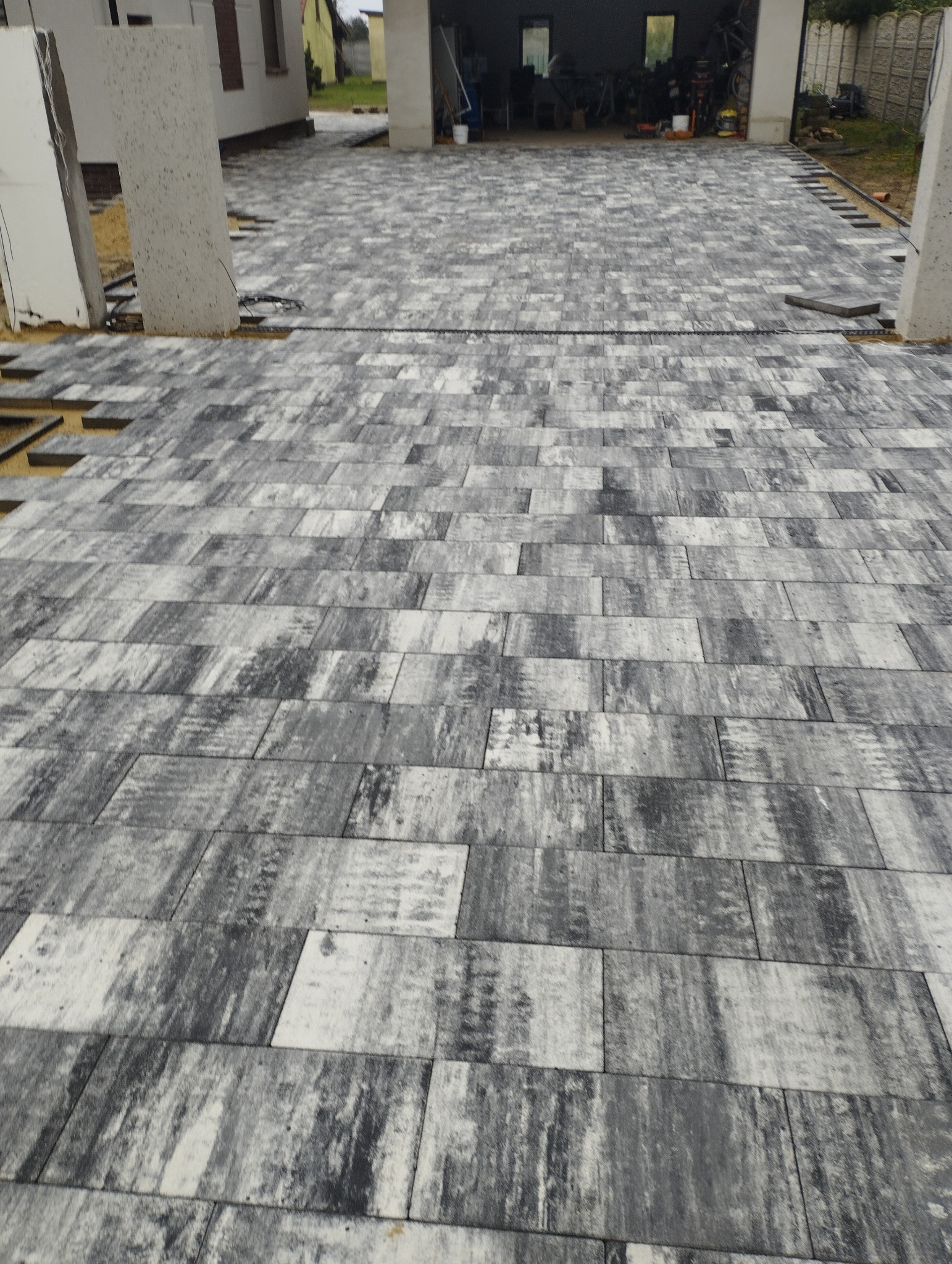 Częściowo ułożona, szara kostka brukowa o prostokątnym kształcie na podjeździe przed domem. Widoczne niezakończone fragmenty układania i elementy konstrukcyjne domu w tle.