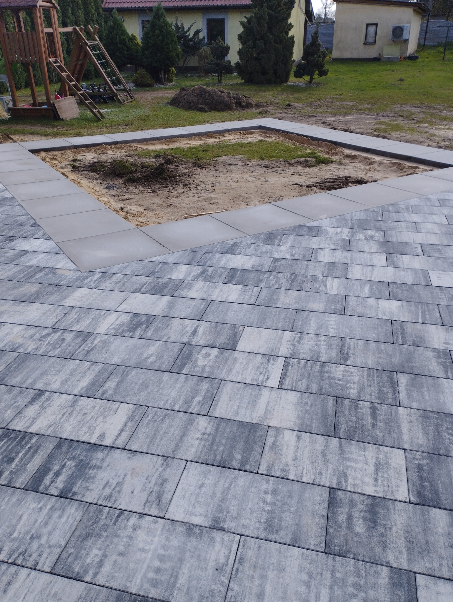 Ułożona szara kostka brukowa z obramowaniem placu zabaw, widok z góry, dom i plac zabaw w tle.