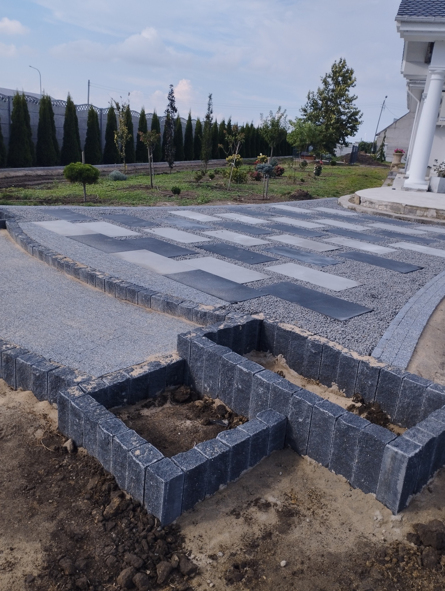 Nowoczesna ścieżka z szarych płyt i grysu prowadząca do domu z białymi kolumnami; w pierwszym planie obrzeże z kostki tworzące rabaty.