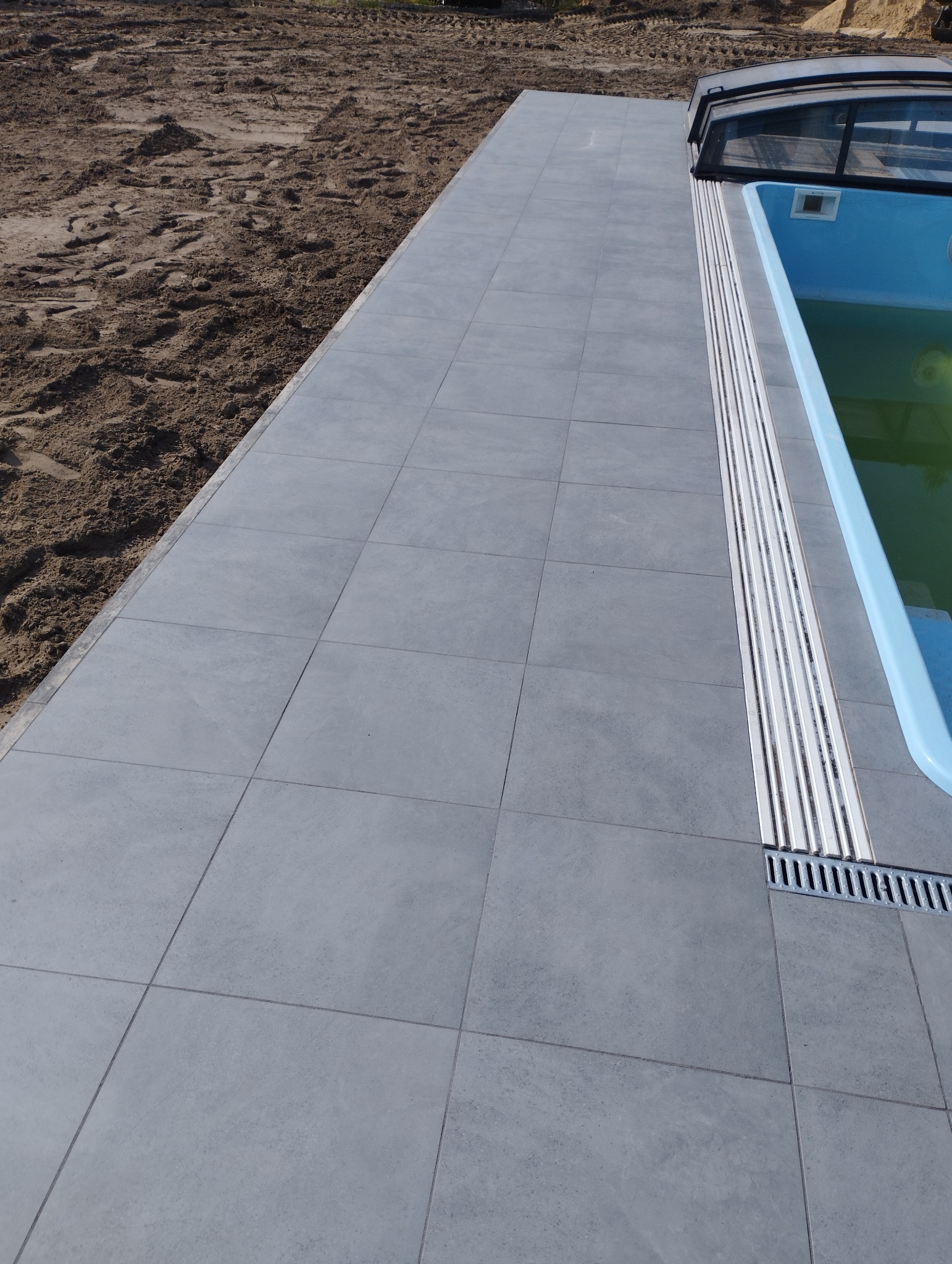 Szara, kwadratowa nawierzchnia z płytek wokół basenu z widocznym odpływem liniowym i przygotowanym terenem pod dalsze prace.