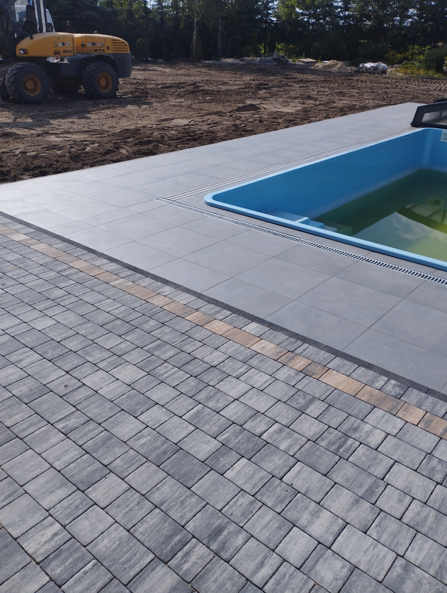 Obszar wokół basenu wyłożony szarą kostką brukową z dekoracyjnym pasem jasnych kostek, w tle żółta koparka i przygotowany teren pod ogród.