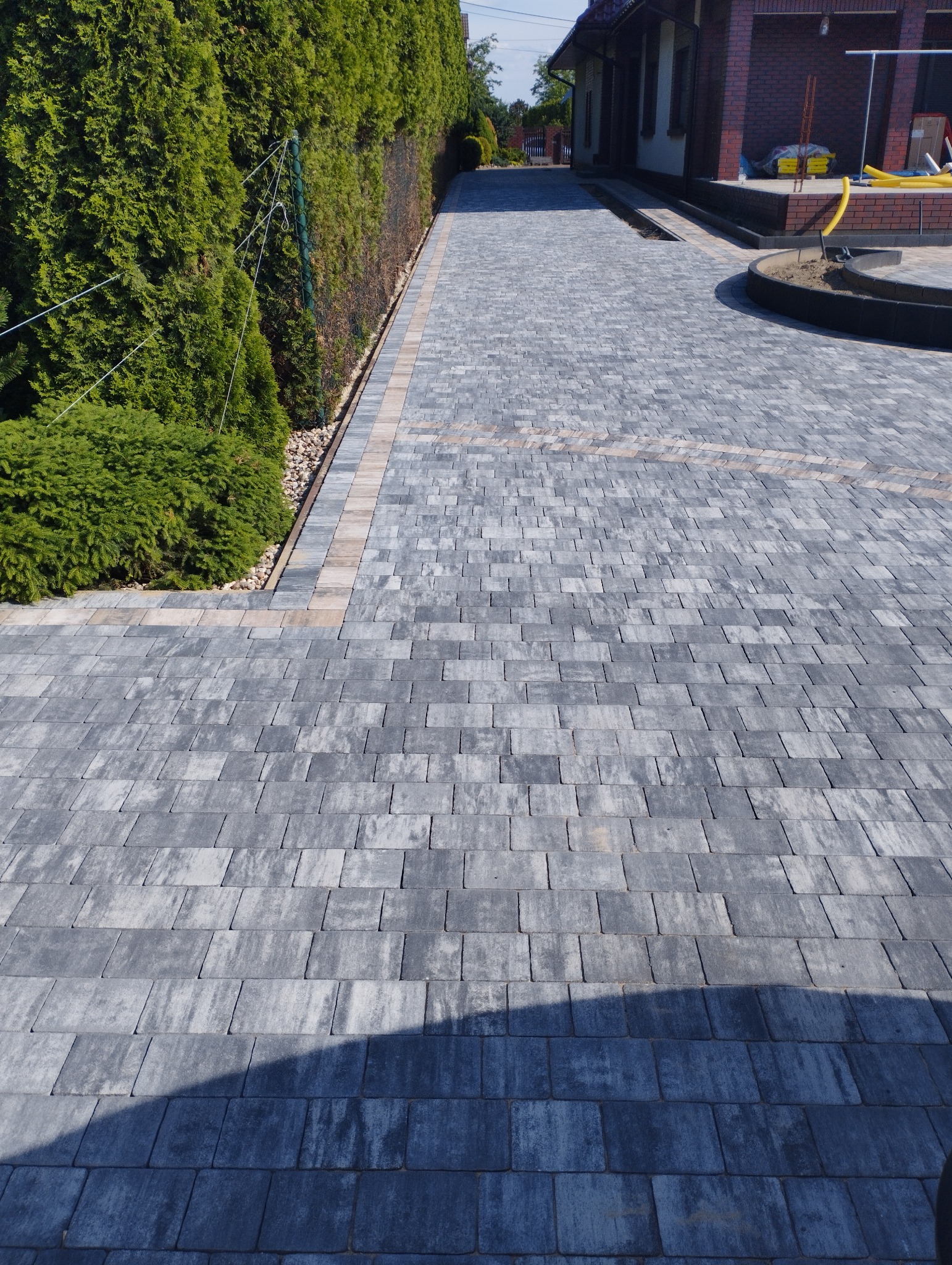 Ułożona, szara kostka brukowa na podjeździe w Kaliszu, z bordiurą z jaśniejszych kostek, obok żywopłot z tui i częściowo wykończony teren wokół domu.