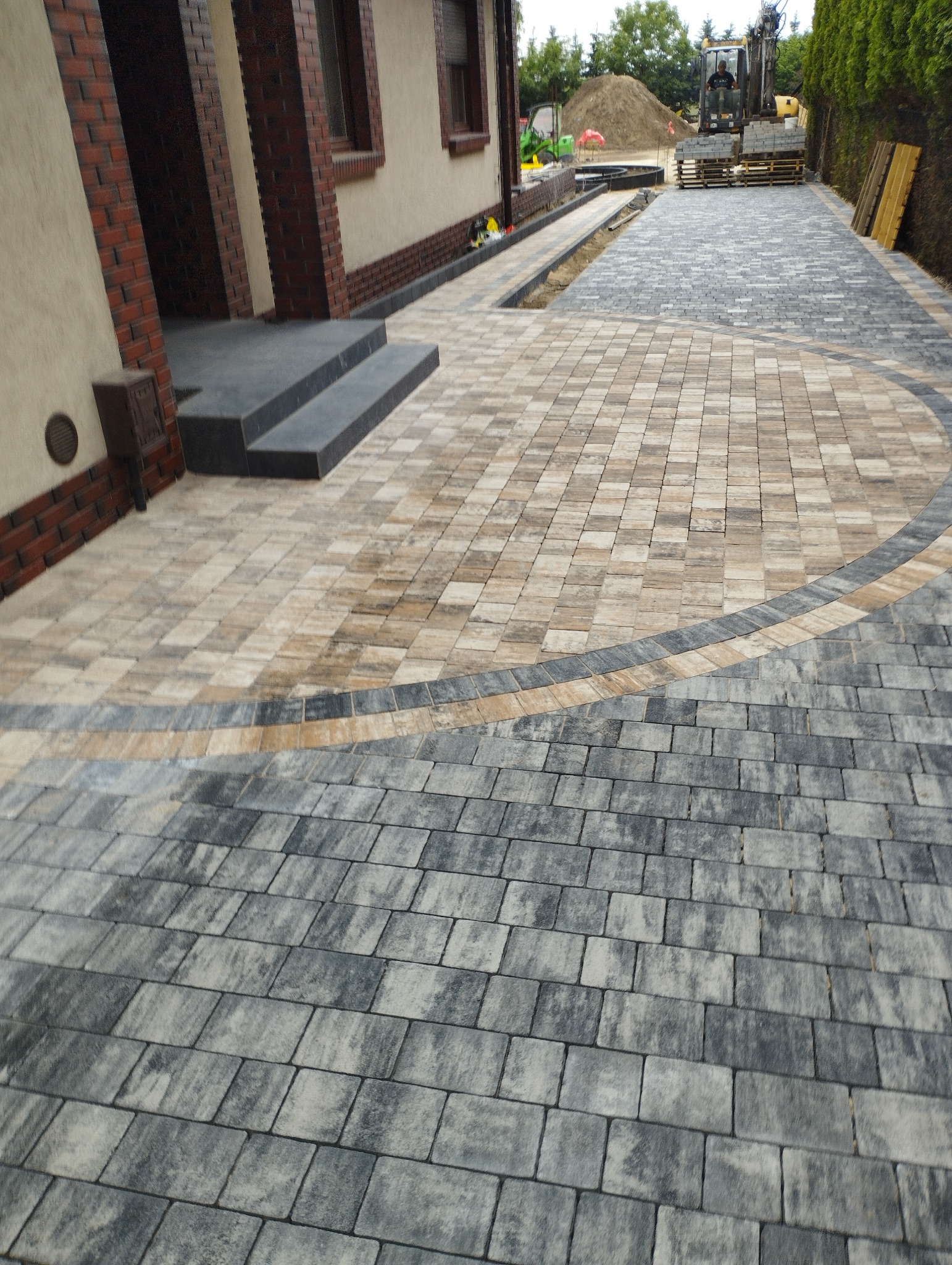 Ułożona kostka brukowa w odcieniach szarości i beżu tworząca wzór na podjeździe, z widocznym domem z ceglanymi elementami i sprzętem budowlanym w tle.
