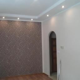 USŁUGI BUDOWLANE A - ZBUD KATARZYNA GRUSZKA - Wykończone wnętrze pokoju z drewnianą podłogą, wzorzystą tapetą na jednej ścianie, białą ścianą z łukowym przejściem do innego pomieszczenia i nowoczesnym oświetleniem sufitowym z halogenami.