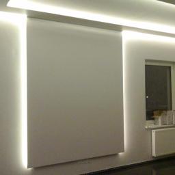 USŁUGI BUDOWLANE A - ZBUD KATARZYNA GRUSZKA - Nowoczesne wnętrze z oświetleniem LED wzdłuż ścian i sufitu, minimalistyczna roleta, otwarte drzwi balkonowe i okno z grzejnikiem.