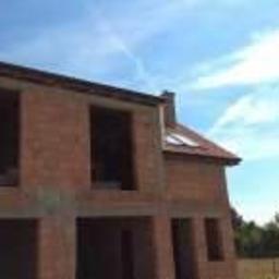 USŁUGI BUDOWLANE A - ZBUD KATARZYNA GRUSZKA - Surowy stan dwukondygnacyjnego domu z czerwonej cegły, widoczne otwory okienne i dach pokryty częściowo czerwoną dachówką, w tle zieleń i błękitne niebo.