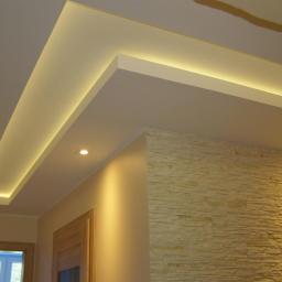 USŁUGI BUDOWLANE A - ZBUD KATARZYNA GRUSZKA - Nowoczesny sufit podwieszany z oświetleniem LED w kształcie litery L, fragment ściany wykończony okładziną kamienną w odcieniach beżu, widoczne drzwi z drewnianą ościeżnicą.