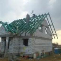 USŁUGI BUDOWLANE A - ZBUD KATARZYNA GRUSZKA - Wznoszenie surowego domu jednorodzinnego z białego bloczka, widoczna konstrukcja dachu z zielonej impregnacji, komin w trakcie budowy, na dachu pracownik w ciemnej odzieży.