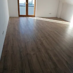 Ukończona podłoga z paneli imitujących ciemne drewno w jasnym, poddaszowym pokoju z białymi ścianami i dwuskrzydłowymi drzwiami balkonowymi.