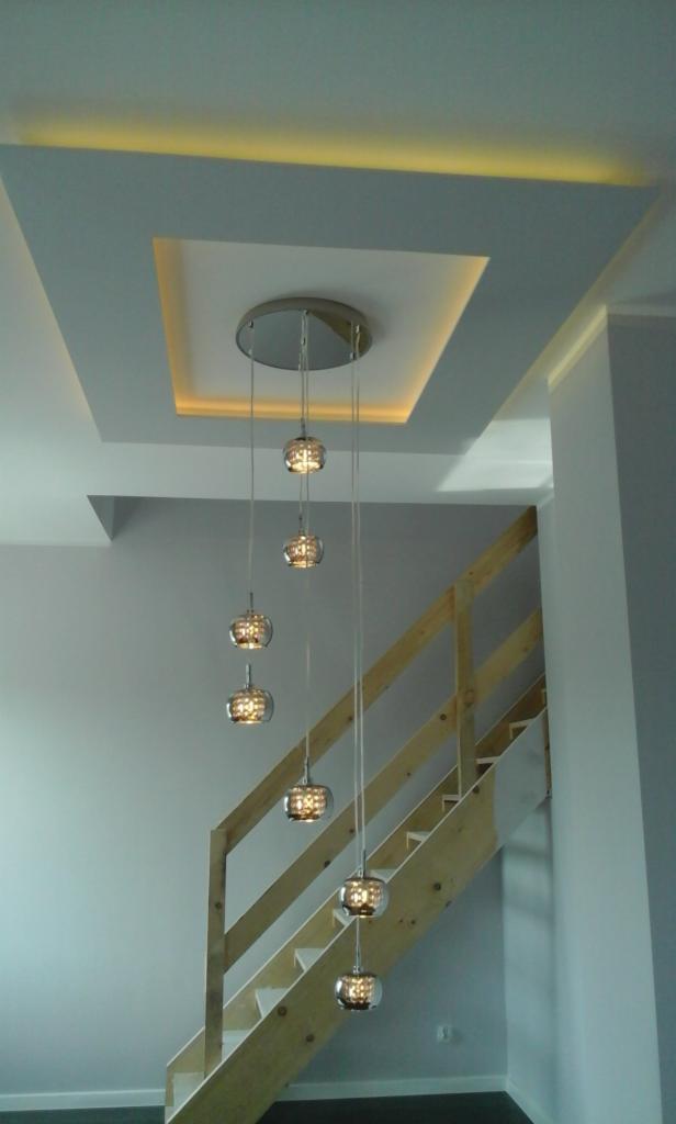 Nowoczesne oświetlenie sufitowe z dekoracyjnymi kulami zwisającymi na tle drewnianych schodów i podświetlanego sufitu podwieszanego.