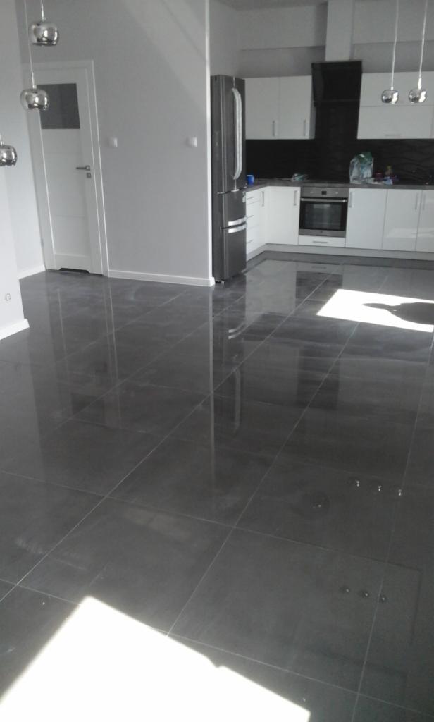 Błyszcząca, ciemnoszara podłoga z płytek w nowym mieszkaniu, widok na białą kuchnię z czarnym panelem, oświetlenie sufitowe.