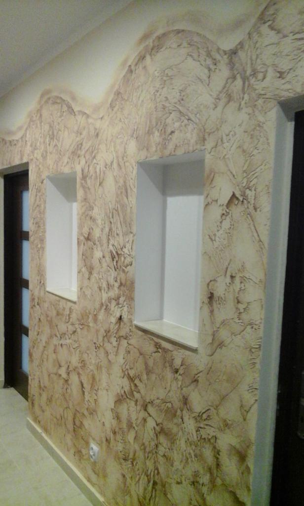 Ściana w korytarzu pokryta dekoracyjnym tynkiem w odcieniach beżu i brązu, z dwoma wnękami pomalowanymi na biało i widocznym fragmentem drzwi po lewej stronie.