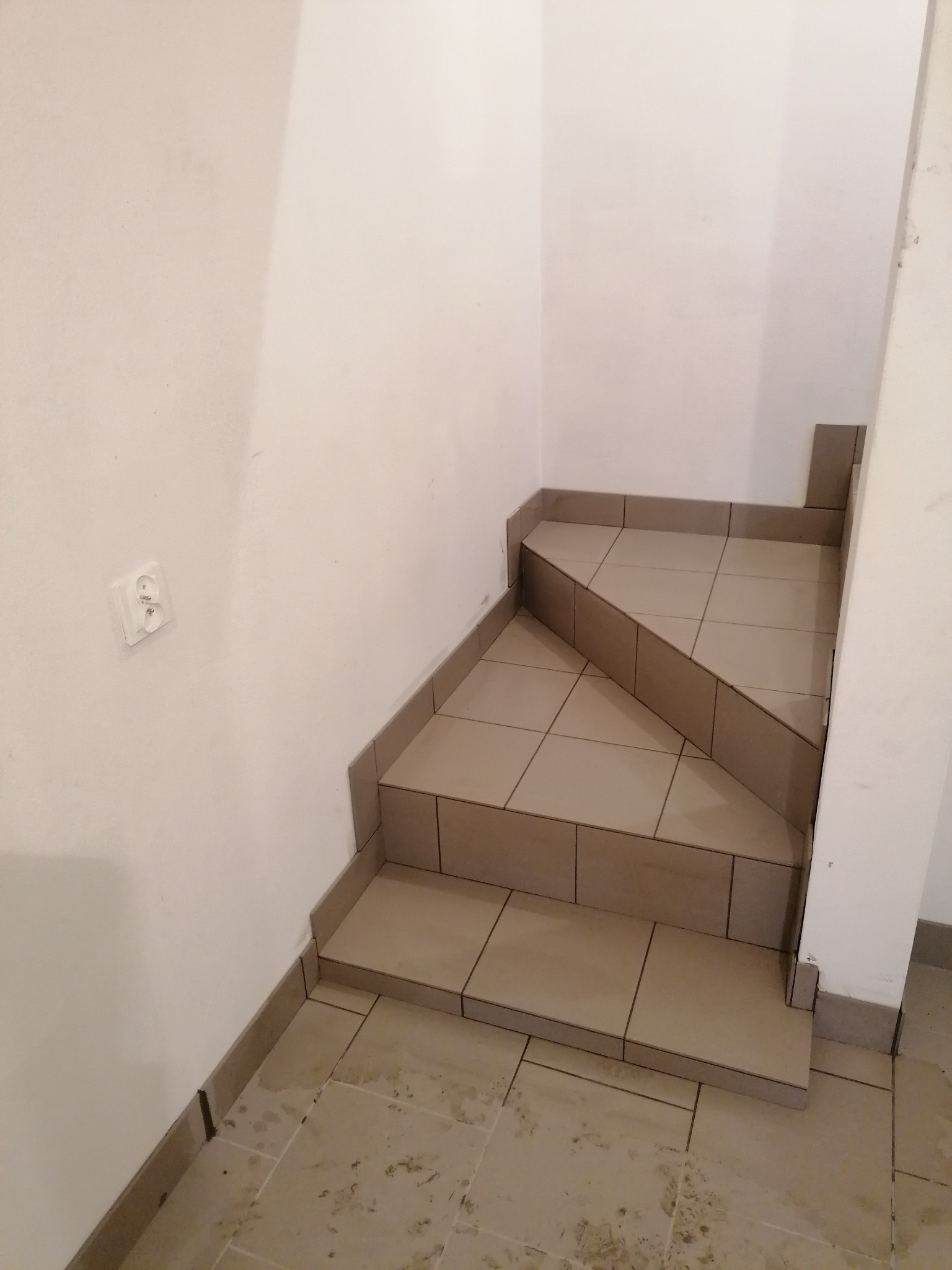 Schody wyłożone beżowymi płytkami ceramicznymi w trakcie prac wykończeniowych, widoczny fragment ściany z gniazdkiem elektrycznym i zabrudzenia na podłodze.