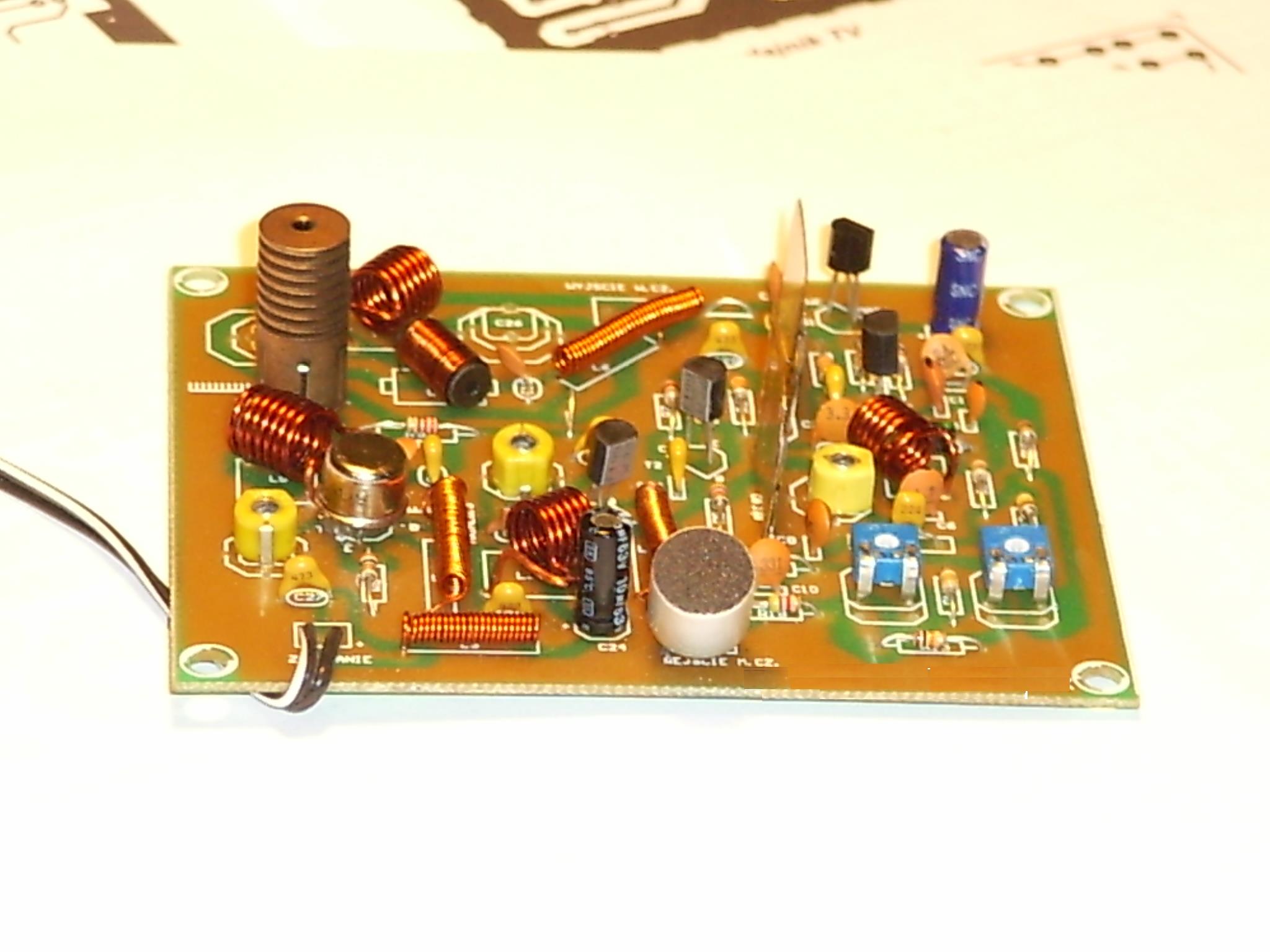 Płytka drukowana z licznymi elementami elektronicznymi, w tym cewkami, kondensatorami i mikrofonem, ułożona na białym tle z fragmentem schematu w tle.