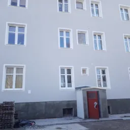 Odnowiony budynek wielorodzinny z szarą elewacją, białymi obramowaniami okien i czerwoną budką przy wejściu, widoczne ułożone deski i ślady po pracach ziemnych.