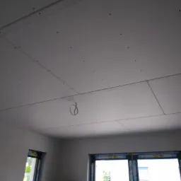 Montaż sufitu z płyt kartonowo-gipsowych w nowym budynku, widoczne łączenia płyt i przewody elektryczne wystające z sufitu, okna z widokiem na zewnątrz.