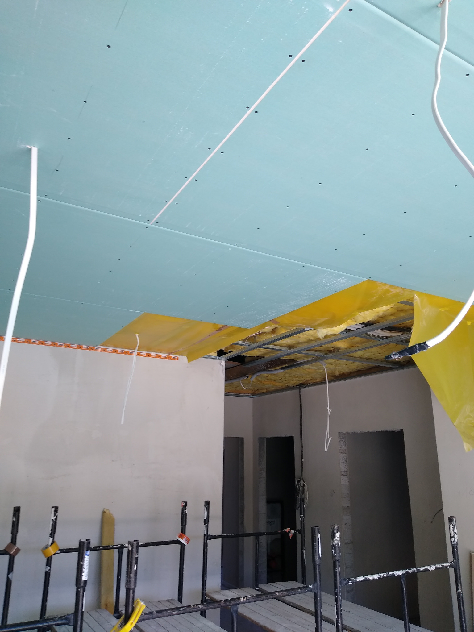 Montaż sufitu podwieszanego: widok na metalową konstrukcję z izolacją, częściowo zakrytą żółtą folią, oraz fragment zamontowanej, jasnoniebieskiej płyty z widocznymi punktami mocowania, z poziomu...