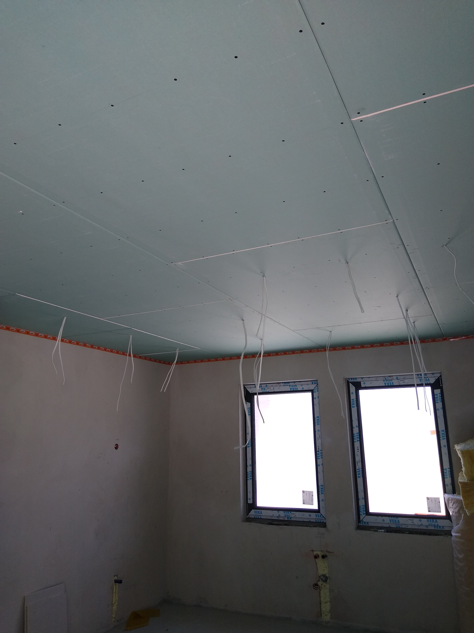 Montaż sufitu z płyt kartonowo-gipsowych, widoczne łączenia płyt, wystające kable elektryczne, dwa okna z ramami VEKA oraz fragment ściany z instalacją hydrauliczną w surowym stanie.