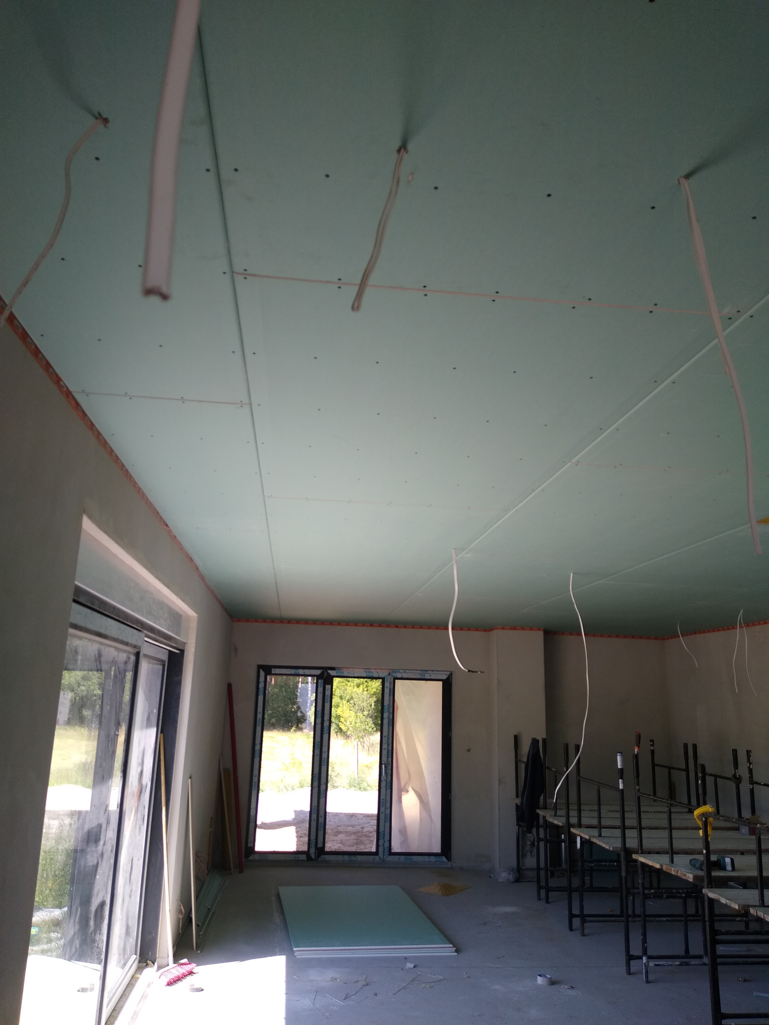 Montaż sufitu z płyty karton gips z widocznymi kablami zwisającymi z konstrukcji, częściowo wykończone ściany i okna w surowym stanie, rusztowanie w tle.