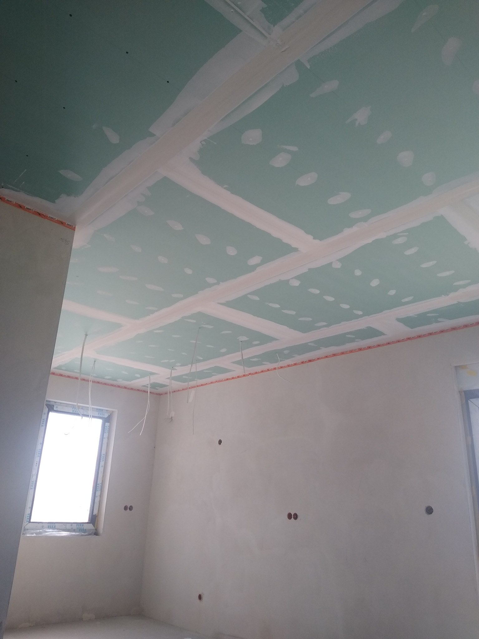 Montaż sufitu z płyt gipsowo-kartonowych na metalowej konstrukcji, widoczne łączenia płyt wypełnione masą szpachlową, kable zwisające z sufitu, surowy stan wykończenia wnętrza.