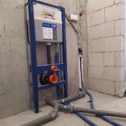 Stelaż podtynkowy Cersanit System z widocznymi przyłączami kanalizacyjnymi z szarego PCV oraz rurami doprowadzającymi wodę w izolacji koloru niebieskiego, zamontowany w surowym stanie...