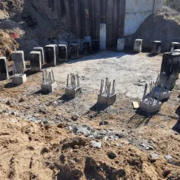 Przygotowanie fundamentów pod budynek: betonowe słupy z wystającymi prętami zbrojeniowymi, ułożone na planie przyszłego budynku, otoczone wykopem w ziemi.