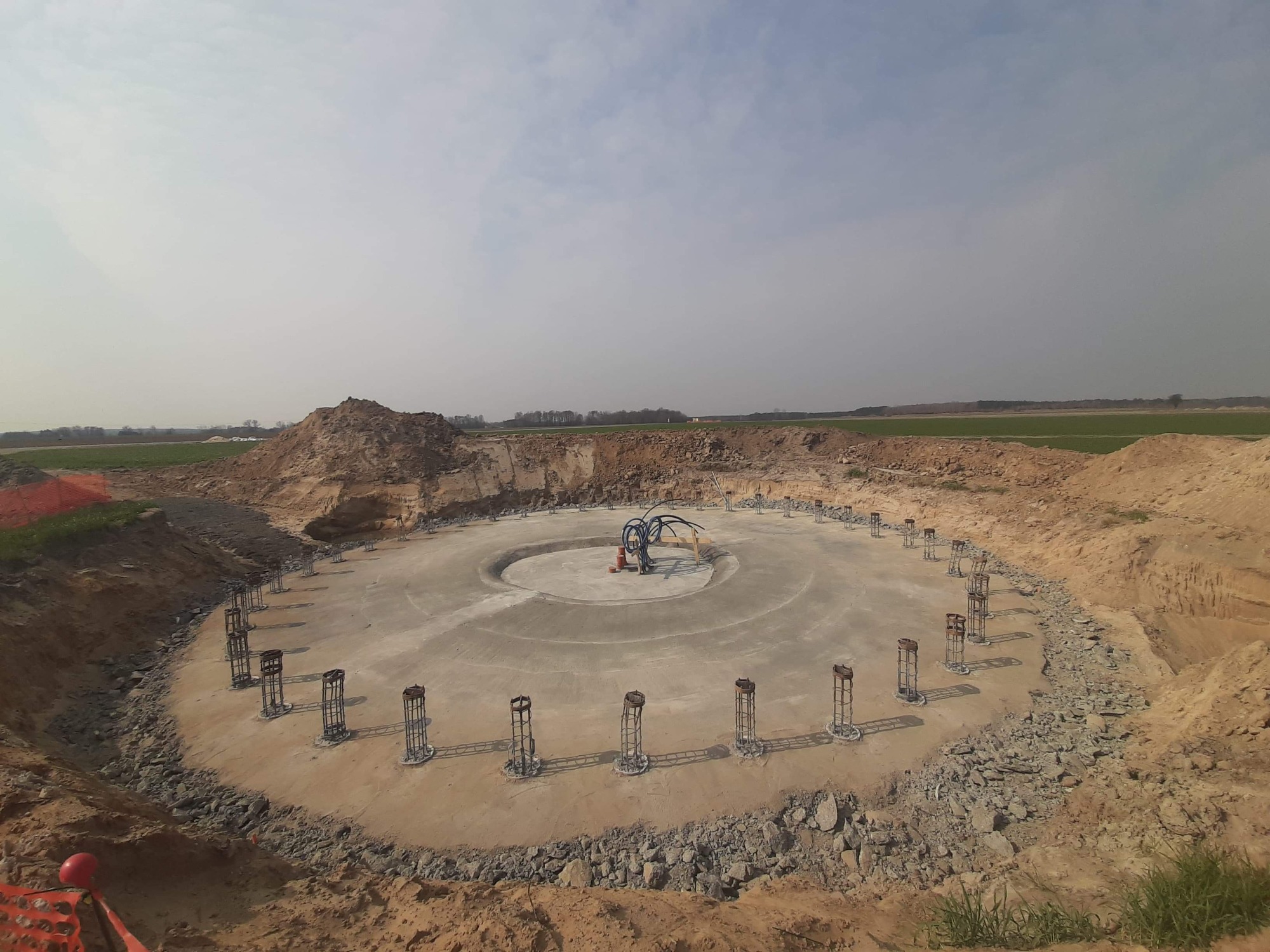 Widok z góry na okrągły fundament z betonu, otoczony ziemią i zbrojonymi słupkami, przygotowany pod konstrukcję, prawdopodobnie wiatrak. Na środku widoczne rury i sprzęt budowlany.