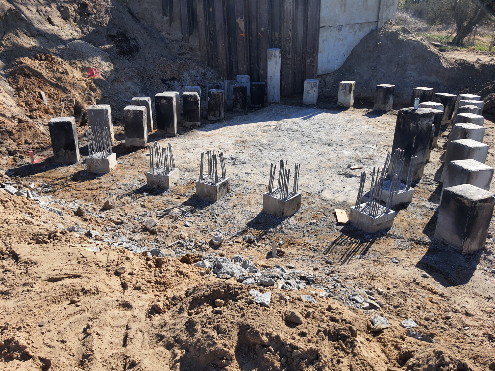 Przygotowanie fundamentów pod budynek: betonowe słupy z wystającymi prętami zbrojeniowymi, ułożone na planie przyszłego budynku, otoczone wykopem w ziemi.