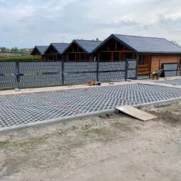 Ułożona szara ażurowa nawierzchnia z obrzeżem z kostki betonowej, widoczne domki letniskowe i ogrodzenie panelowe w tle.