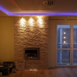 Biel-Bud - Salon z kominkiem obłożonym dekoracyjną, kamienną płytką w odcieniach beżu, oświetlonym trzema punktowymi lampami. Sufit z niebieskim oświetleniem LED.
