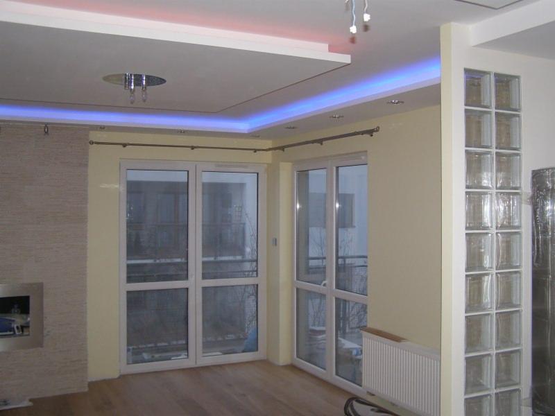 Jasne wnętrze z oknami balkonowymi i ścianką z luksferów, oświetlone listwami LED w suficie podwieszanym. Widoczny grzejnik i fragment podłogi.